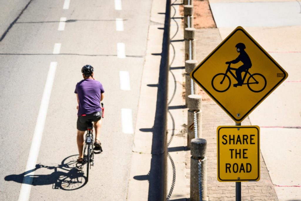 UW-Madison bicyclist riding on the road in bike lane near a road sign that contains a bicyclist symbol and "SHARE THE ROAD"
