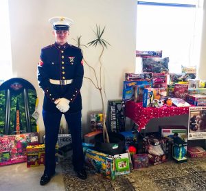 person in military uniform stands with hands clasped in white gloves and wearing a hat in front of a tall plant and a very large table of toys and wrapped gifts that spill out onto the floor, all in front of a large illuminated window
