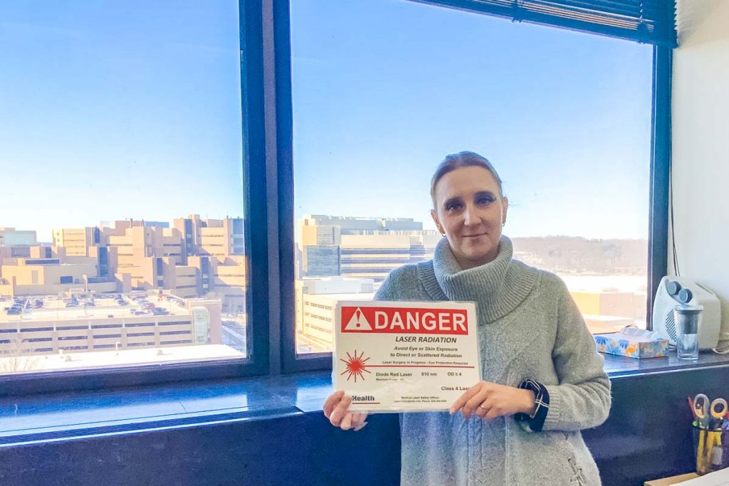 Woman holds "DANGER" sign regarding laser safety while standing in front of a window view of campus