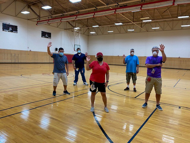 five individuals wearing face coverings are standing in a gymnasium and waving