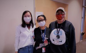 3 people wearing masks and holiday clothing stand together in a hallway