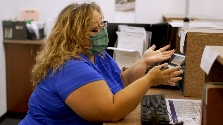 woman wearing a face mask sits at a desk and gestures with hands as if in conversation with someone outside the frame