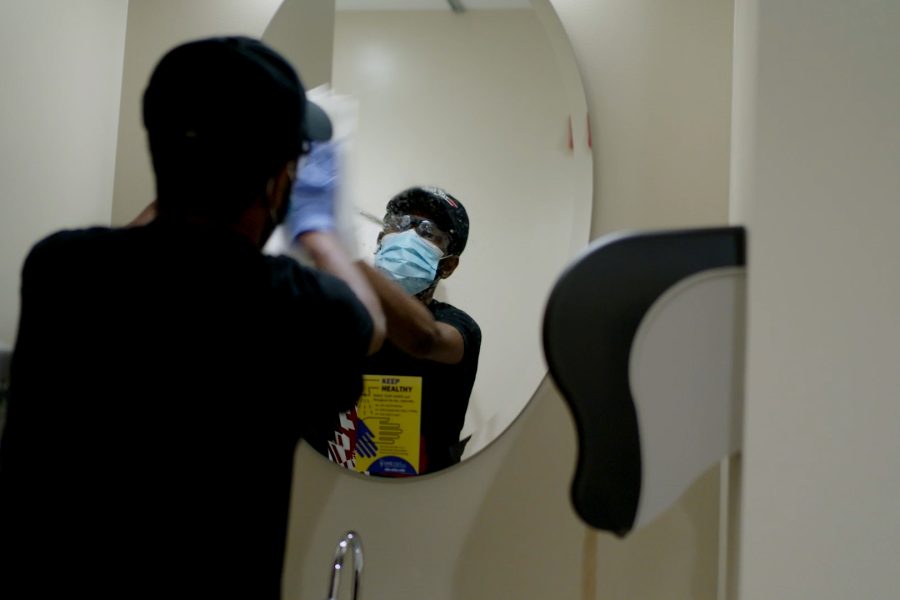 custodian cleaning mirror while looking at reflection while wearing a disposable face mask