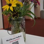 flowers in a jar on a table with white tablecloth. Next to it sits a tent card that reads "We want your feedback. Answer 5 quick questions:" with a QR code beneath it.