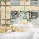 A Grounds crew member drives a green and yellow piece of snow removal equipment that brushes snow off a wide sidewalk in front of a campus building.
