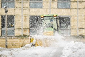 A Grounds crew member drives a green and yellow piece of snow removal equipment that moves snow off a wide sidewalk in front of a campus building.