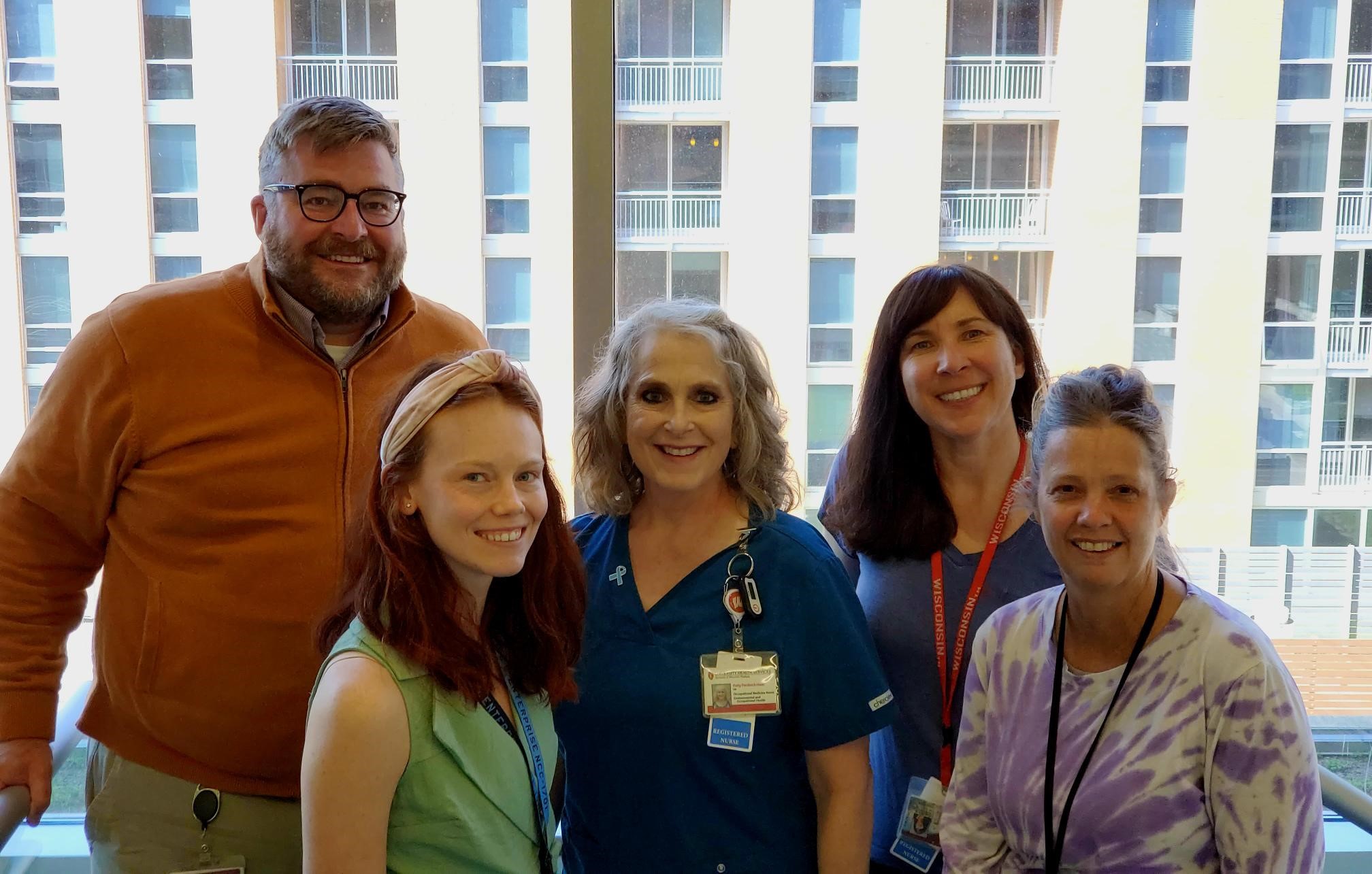 Five people standing in front of a lit window in the daytime wearing nurses scrubs, business casual, tie die. All peple are wearing ID badges and smiling